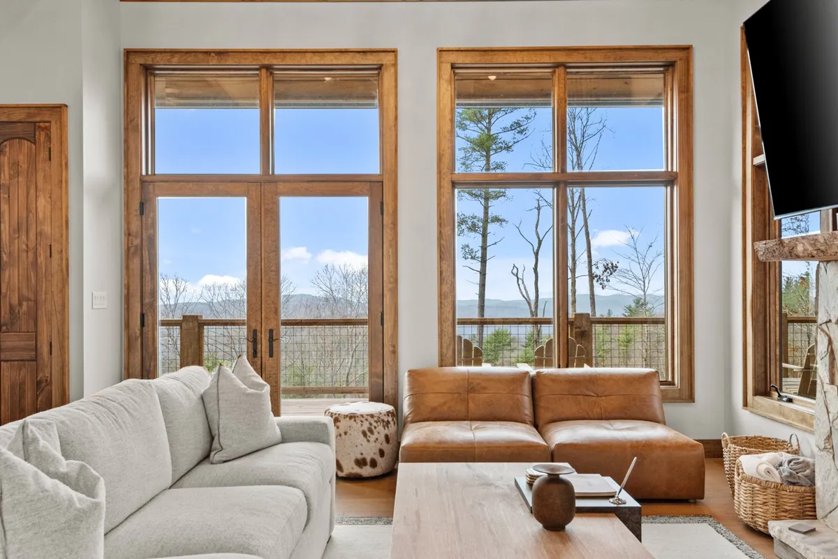 This image features a cozy living area with a large stone fireplace, leather sofa, wooden coffee table, and expansive windows that frame beautiful mountain views. The space is bathed in natural light, creating an inviting setting for gatherings, conversation, and relaxation.