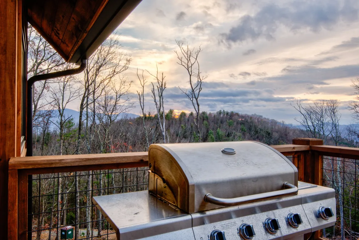 A grill setup on an outdoor patio framed by trees and a spectacular mountain view. The area is designed for barbecuing, entertaining guests, and enjoying sunsets in a scenic natural setting.
