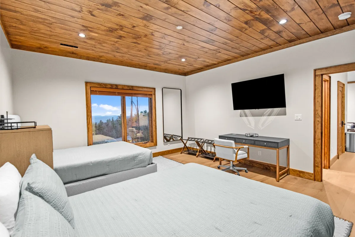 This interior image shows a spacious bedroom featuring two beds with plush linens, a large window offering serene views, a flat-screen TV mounted for entertainment, and a dedicated desk and chair for work or study. Natural light fills the space, enhancing its inviting and peaceful ambiance.