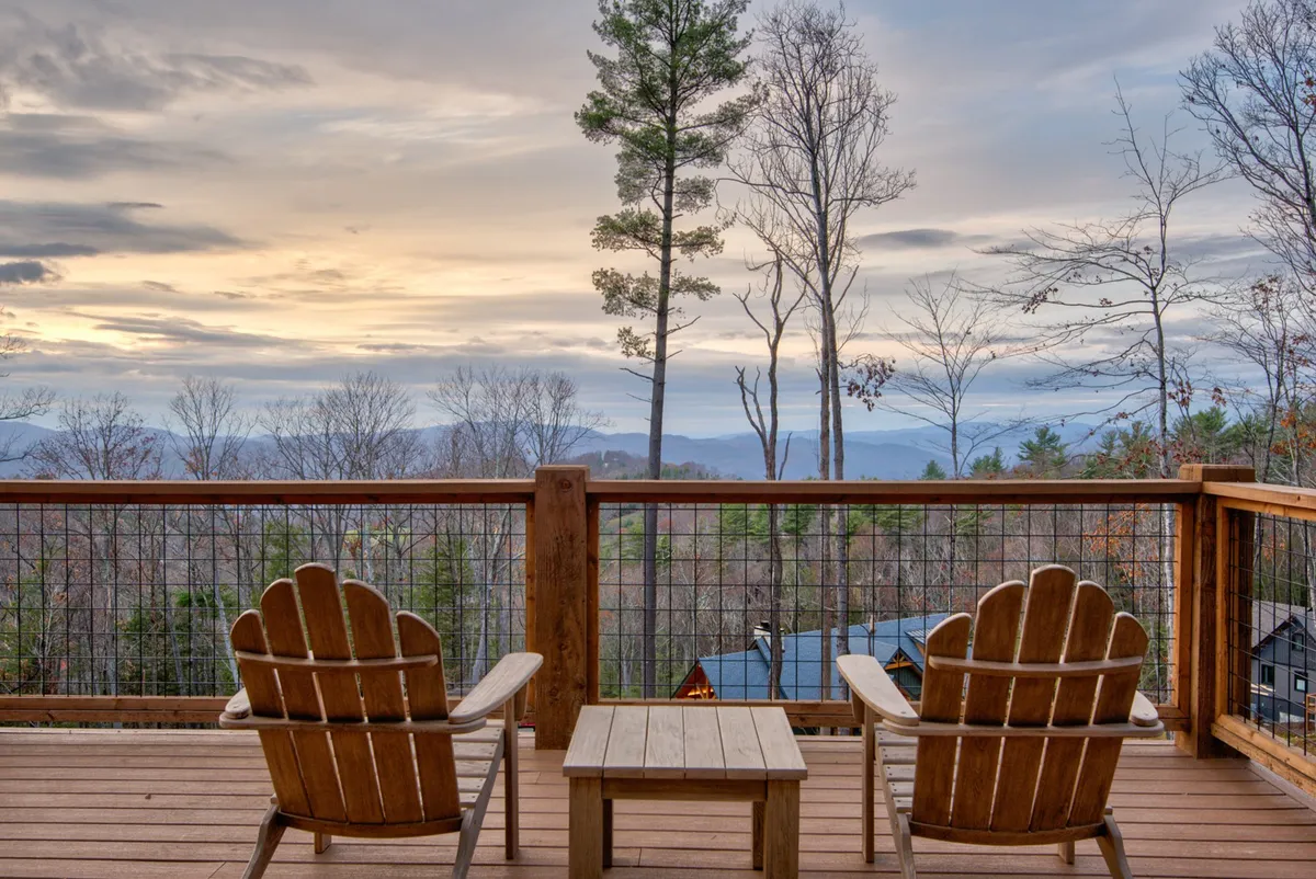 A tranquil wooden deck with Adirondack chairs set out to face sweeping mountain views. The deck is surrounded by trees and overlooks a breathtaking sunset sky, inviting moments of contemplation or socializing in nature's embrace.
