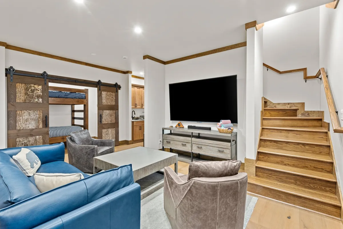 A lower-level recreation room is depicted, complete with a blue sofa, bunk bed, television, and wooden stairs. The setup suggests a versatile space ideal for movie nights, game sessions, and family fun, complementing the main living area.