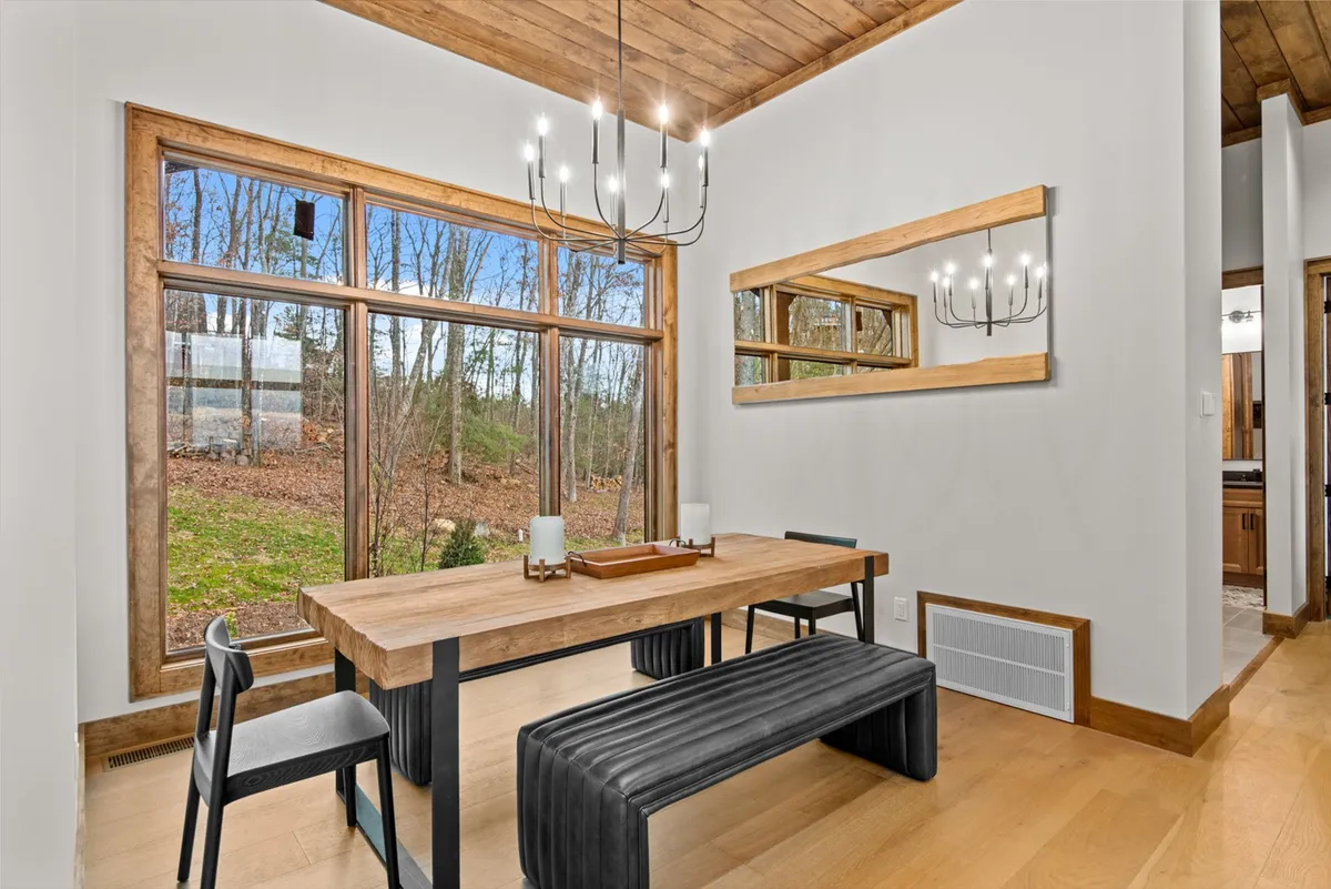 A cozy dining area featuring a large wooden table, bench seating, a stylish chandelier, and floor-to-ceiling windows offering scenic views. The space is set for gatherings, be it intimate family brunches or celebratory dinners, all set against a backdrop of natural beauty.