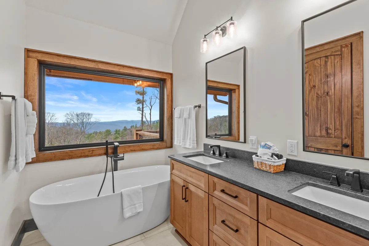 A spa-inspired bathroom featuring a freestanding soaking tub positioned in front of a large window with scenic views, dual sinks, and elegant wooden cabinetry. The space emphasizes relaxation and a boutique hotel feel.