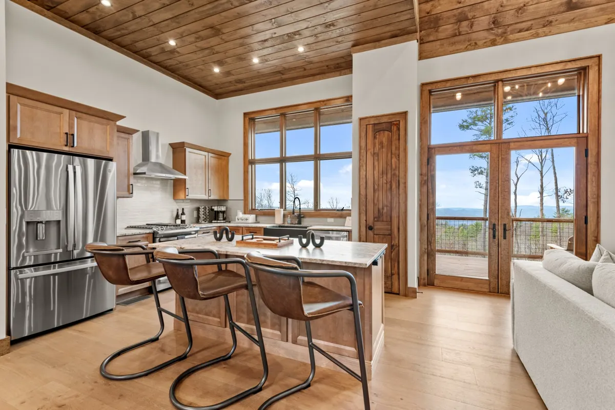 A warm, inviting kitchen with a large island, modern appliances, wooden ceiling, expansive windows, and bar stools. The setup emphasizes the social heart of the home, where cooking and gathering seamlessly blend together. Natural light fills the space, creating a cheerful ambiance ideal for both daily meals and festive occasions.