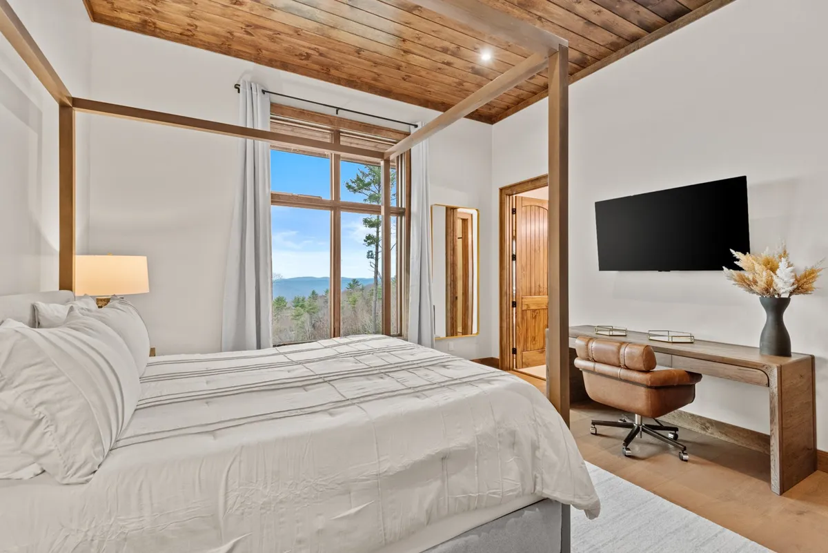 A tranquil bedroom featuring a four-poster bed with comfortable linens, a large window framing a scenic view, a wooden desk and chair for work-from-home needs, and a wall-mounted flat-screen TV for entertainment. The color palette and furnishings evoke a sense of calm and rejuvenation.