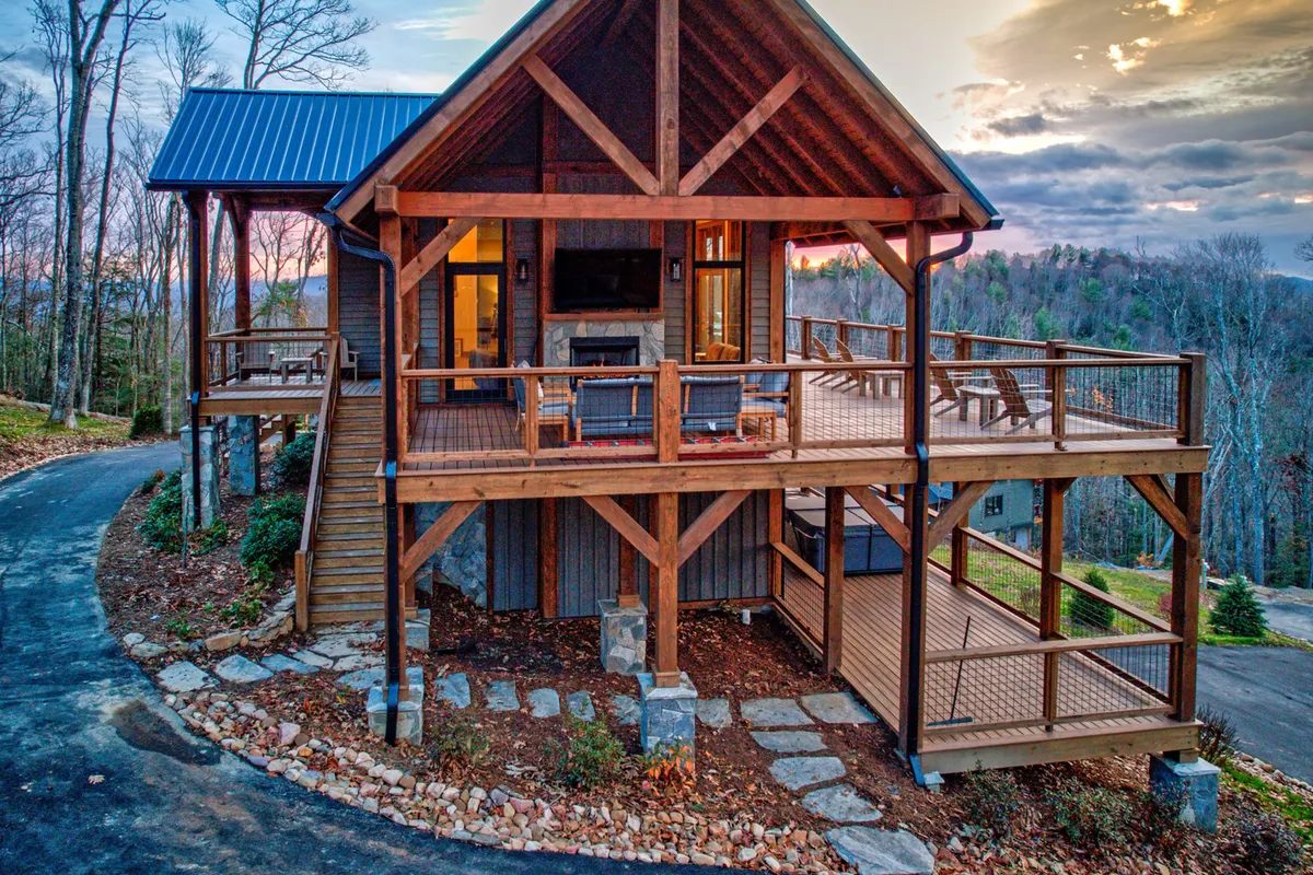 The image shows a spacious single-family mountain home with a large wooden deck overlooking sweeping mountain views. The deck features comfortable seating for outdoor dining, a stone pathway leading to a cozy fire pit, and large windows that invite in natural light. The porch is ideal for relaxing, surrounded by tranquil forested scenery, and the overall setting is peaceful and inviting.
