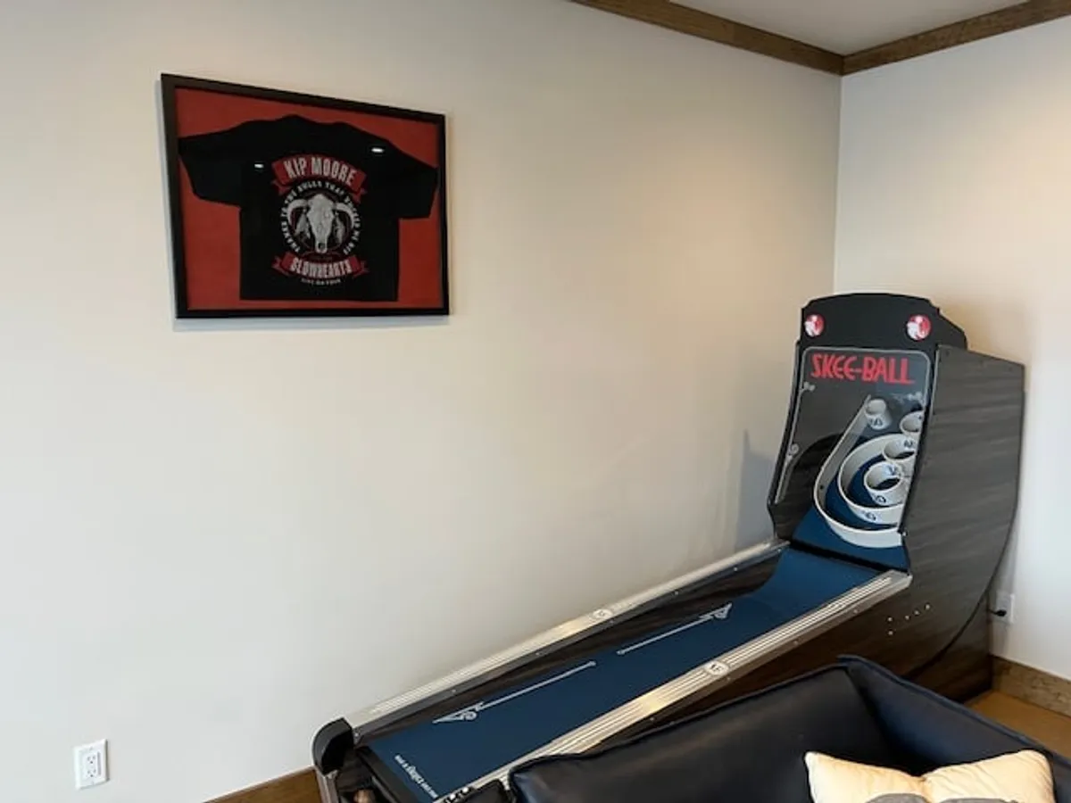 The image spotlights a Skee-Ball machine and a framed t-shirt, emphasizing the playful side of the home. The setup is ready for friendly competition and game nights, highlighting a unique amenity mentioned in the description.